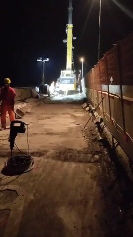 A construction site at night. In the foreground, a heavy-duty jackhammer lies on a concrete surface. Behind it, a person in an orange safety suit and a yellow hard hat stands on the left side of the frame, facing away from the camera. Further ahead, a large yellow crane is positioned, its boom extended upwards. Several construction workers in orange safety suits are visible near the crane, some appearing to be engaged in work around the vehicle. The environment is dimly lit by artificial lights, and orange construction barriers line the right side of the frame. The ground appears to be a mixture of concrete and dirt, with debris scattered around.