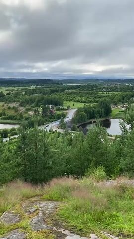 This media file provides a high-angle view of a rural landscape under a cloudy sky, likely taken from a hilltop overlooking a valley. The scene is dominated by lush green trees, fields, and bodies of water, suggesting a temperate climate. A paved road with a bridge crosses through the middle of the frame, with several cars visible, indicating the presence of some transportation activity. A few scattered houses and buildings are nestled among the trees, hinting at a sparsely populated area. The foreground features rocks and tall grass, common in elevated, natural settings. The overall impression is one of serene, natural beauty, with a touch of human presence through the road and vehicles. The cloudy sky suggests it could be overcast or nearing dusk or dawn, but without more definitive cues, pinpointing the exact time of day is difficult. The location context provided places this scene in Lakhdenkiulya, Russia, which aligns with the typical scenery of such regions. There is no visible text within the media file.