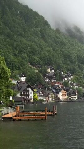 A low-angle video pans across a lake to show a village nestled on the side of a mountain covered in trees. The village is made up of several houses with dark roofs and some white walls. There is a small wooden dock with railings on the left side of the frame. The sky is overcast and foggy. The water is dark and appears choppy.