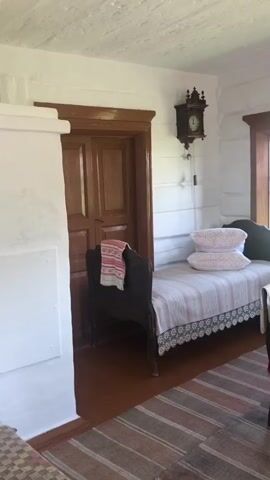 The video pans across a rustic room with white-washed walls and a wooden ceiling. On the left, a white stove-like structure stands next to a dark wooden door with two panels. A folded towel with red and white stripes is draped over a dark wooden bench placed in front of the door. To the right of the door, a narrow wooden bed is covered with a striped pink and white blanket adorned with a white lace trim. Two pillows, also pink and white, rest on the bed. A dark wooden pendulum clock hangs on the wall above the bed. Further to the right, a round table is covered with a red tablecloth featuring a white floral pattern and fringed edges. Two chairs with light-colored seats are pulled up to the table. A glass oil lamp with a white base sits on the table. Two windows with wooden frames are visible on the right wall, looking out onto an outdoor scene with wooden structures. A dark wooden bench is positioned beneath the windows, between the table and a corner of the room. A striped rug with red, pink, and beige stripes covers the wooden floor. The room appears to be part of a traditional dwelling.