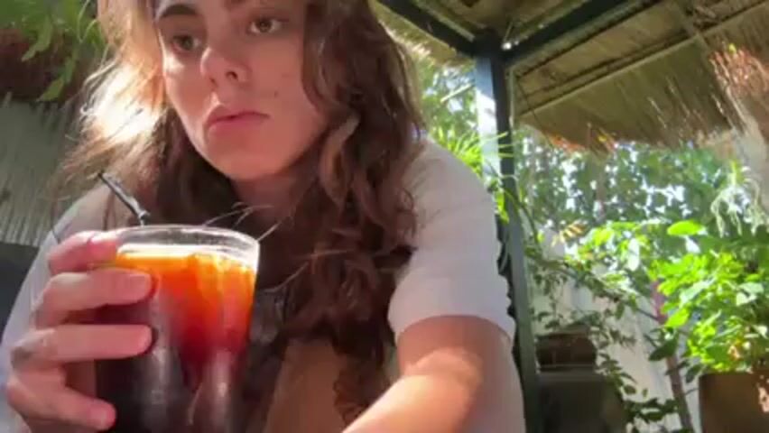 A young woman with curly brown hair is sitting outside, and she is pouring a creamy liquid from a small glass into a larger glass containing a dark liquid, presumably coffee. The larger glass has a black straw in it. The background includes plants and a thatched roof structure.