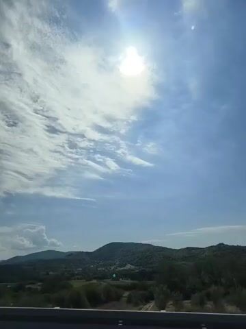 The video shows a bright, sunny day with a partly cloudy sky. The sun is high and bright, with some wispy clouds scattered around it. Below the sky is a landscape of rolling green hills covered in trees. In the foreground, there is a railing that suggests the video was taken from a bridge or an elevated road. There are no people or animals visible in the video. The overall impression is one of a serene and beautiful natural landscape.