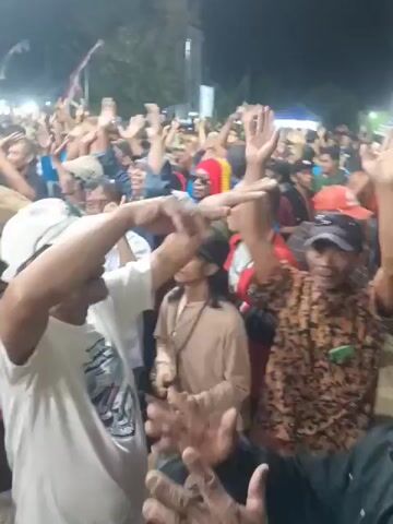 The video shows a crowded outdoor gathering at night. Many people are visible, with some in the foreground and others further back. The atmosphere appears festive, as many individuals have their arms raised in the air. Some people are wearing hats and casual clothing, like t-shirts and patterned shirts. Lights are visible in the background, suggesting a temporary setup or stage area. The ground appears to be dirt or gravel. The individuals in the foreground are focused on something happening in front of them, with expressions of excitement and engagement. The overall scene suggests a public event, possibly a concert or rally, with enthusiastic participation from the attendees.
