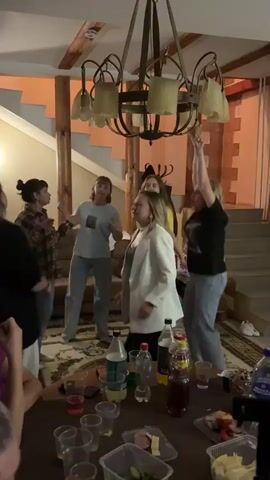A group of people, mostly women, are dancing in a room. They are dressed casually, in jeans and t-shirts. There is a table in the foreground with drinks and food on it. The room has a rustic feel, with wooden beams and a large chandelier. The lighting is dim, and the atmosphere appears to be festive. Some of the people are smiling and laughing, and their body language suggests they are enjoying themselves.