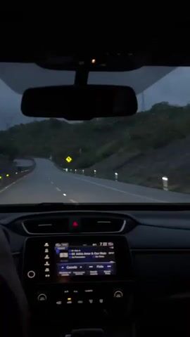 A first-person point of view video shows a person driving a car on a winding road at dusk or dawn. The car is on a highway with two lanes visible, one going in each direction. There are green hills on both sides of the road. The road has yellow lane markers and white reflective markers along the shoulder. A yellow road sign with a black curved arrow is visible on the right side of the road. The car's dashboard and infotainment system are visible at the bottom of the frame, showing music playing. The car's headlights illuminate the road ahead. The sky is overcast and dim.
