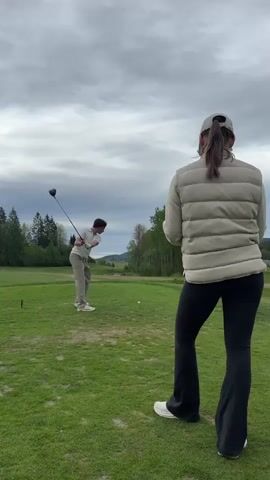 The video shows a man and a woman at a golf course on an overcast day. The man is wearing a white shirt and khaki pants. He swings a golf club and hits a golf ball. The woman is wearing a light-colored vest, black pants, and a baseball cap. She is standing and watching the man. In the background, there are trees and hills.
