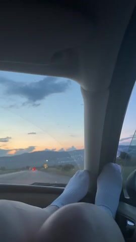 A first-person perspective video shows a person's legs and feet, wearing white socks, propped up in the passenger seat of a car. The legs and feet are close to the camera. Through the car's windshield, a sunset is visible with mountains in the distance and a road with cars driving. The sky is blue with clouds tinged with orange and yellow from the setting sun. The interior of the car is dark.