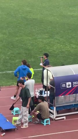 A video shows an individual standing on a track at a soccer field, holding a microphone, possibly making announcements or speaking to the players on the field. He is wearing a navy blue polo shirt and black pants. Other people are standing or sitting near the sideline, while soccer players are visible on the field in the background. A sign with "CFA 2024" is displayed beside a team bench.