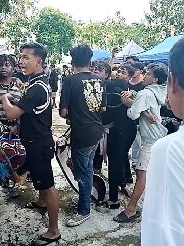 The video shows a crowd of men in what appears to be an outdoor setting. Many are wearing black t-shirts with white and gold logos on the back. One man is wearing a light blue t-shirt with writing on the back. There are some tents and trees visible in the background. The men seem to be engaged in a tense discussion, with some individuals appearing to restrain others. The atmosphere seems potentially volatile.