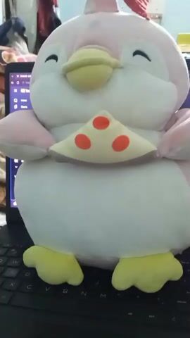A stuffed animal resembling a pink and white chicken with yellow feet and a yellow beak is positioned on a black keyboard. A small, triangular slice of pizza, light yellow with three red circles, is attached to the front of the stuffed animal. The stuffed animal's eyes are closed, and its head is tilted slightly. In the background, a laptop screen is visible, showing a webpage with text and graphics. The environment appears to be indoors, with soft lighting and some blurry items in the background.