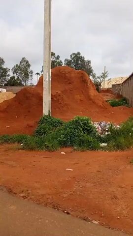 The video shows a construction site with piles of sand and gravel. In the background, there is a building with a green roof, likely a gas station, and a wall. Trees are visible behind the wall. The ground is covered in red dirt, and there are some puddles and debris. The sky is cloudy. The camera pans across the scene, showing different angles of the construction site. There are no people visible in the video.