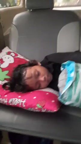 A young person is sleeping on the back seat of a car. They are lying on a pink Hello Kitty pillow with a strawberry. There's a striped blue and green bag next to them. The car seat is gray. The car is moving, as can be seen through the window in the background.