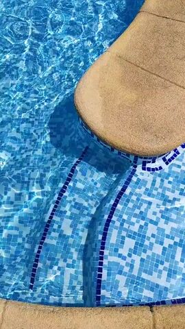 A close-up shot of the edge of a swimming pool shows blue tiles and a tan curved step leading into the water. The water is clear and reflects light, creating ripples and shadows on the tiled steps.