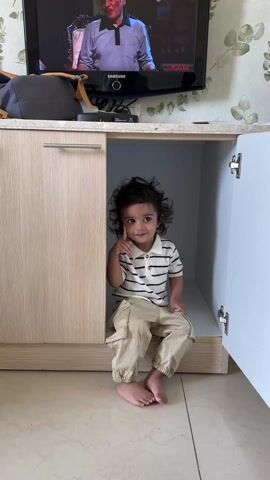 A small child sits inside a cupboard under a television. The child has dark, curly hair and is wearing a striped collared shirt and khaki pants. The child is barefoot and sits on a shelf inside the cupboard, looking directly at the camera. The cupboard has a light-colored wood grain finish and one door is open. The television is mounted on the wall above the cupboard, displaying various scenes, including what appears to be a show with actors. The wall behind the television is covered in a floral patterned wallpaper. A gray bag sits on top of the cupboard to the left of the television. The floor is a light-colored tile.