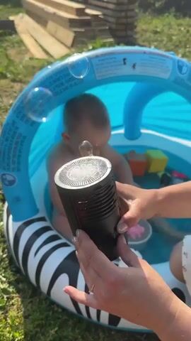 The video shows a baby in a blue and black inflatable pool with a canopy. A person's hands are holding a black bubble machine, which is producing bubbles. The baby is looking at the bubble machine. The pool is in a grassy area, with stacks of lumber in the background. The scene appears to be outdoors on a sunny day.
