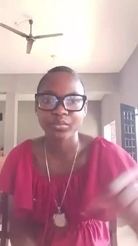 A young Black woman with glasses and a low braided ponytail, wearing a red blouse and a necklace with a Hello Kitty pendant, makes hand gestures while looking at the camera. She is indoors, with a ceiling fan visible above her. The background includes neutral-colored walls and a glimpse of a doorway, suggesting a living or dining area. The lighting appears bright, and the video's perspective is from a slightly low angle. She appears to be giving a message and uses expressive movements.