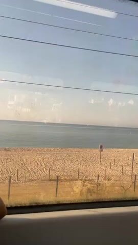 The video shows a view from inside a train, looking out the window at a beach. The beach is sandy with a few wooden posts and some vegetation. The ocean is visible in the background, under a clear sky. A person on a bicycle rides along a path parallel to the beach. The window is slightly dirty, and the video appears to be taken during the daytime.