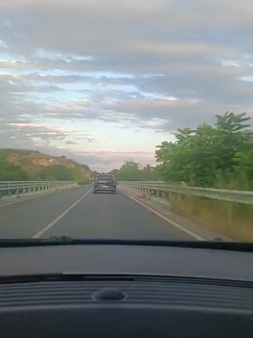 A rear-view video captures a journey along a highway. The camera is positioned inside a vehicle, looking forward. A black SUV is visible ahead on the road, its taillights illuminated. The highway is bordered by guardrails and vegetation. The sky above is filled with clouds, indicating either early morning or late afternoon light. The road appears to be in good condition, with clear lane markings. There are no other vehicles or individuals clearly discernible in the immediate vicinity beyond the SUV and the driver's perspective.