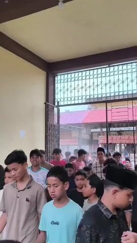 A group of young boys, some wearing traditional Islamic skullcaps, are standing in a line, facing forward and looking towards the camera. The boys appear to be outdoors, possibly in a courtyard or a covered walkway, with a building visible in the background. The building has a yellow wall and a brown trim around a doorway or opening, through which more people can be seen. The atmosphere seems orderly, with the boys positioned in a formation, suggesting they might be waiting for an event or activity.