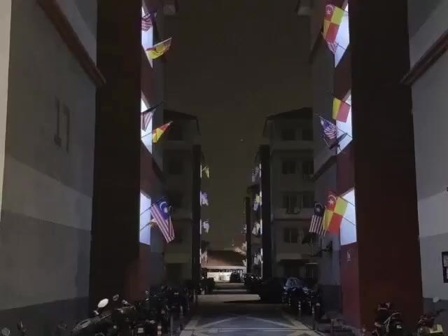 A drone flies through an alleyway between two apartment buildings at night. The buildings are adorned with flags, many of which are Malaysian flags. The flags are illuminated by lights from within the building windows. Motorcycles are parked on either side of the alley. The drone moves forward, passing by multiple windows on each building, each with flags hanging from it. The drone continues to fly down the alley until it reaches the end.