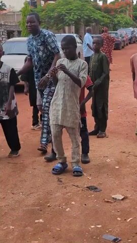 A medium shot video shows a group of men, women, and children on a dirt road. A tall man in a blue and white patterned outfit with brown sandals and two children, one in a red shirt and jeans and another in a cream-colored top and pants with blue slides, are walking towards the left of the frame. The tall man is smiling and dancing while the children are looking at the camera. Another child in a green top and pants with black sandals is standing on the right. In the background, other people are visible, and several cars are parked along the side of the road. The road is unpaved and a few pieces of trash are visible on the ground.