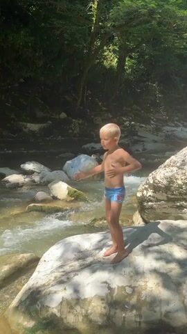 A young boy and a baby girl are by a river. The boy is standing on a rock and looking at the camera. The baby girl is standing on the ground and crying. The river is flowing and there are rocks and trees in the background.