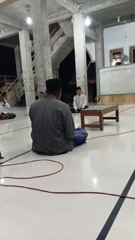 The video shows a group of men sitting on the floor in a room with a concrete ceiling and columns. The men are all wearing traditional Islamic clothing. Some of them are wearing white shirts and pants, while others are wearing more casual clothing. The men appear to be engaged in a religious activity, as some of them are bowing their heads in prayer. There is a whiteboard in the background, which may be used for educational purposes. The room is dimly lit, with lights on the ceiling providing illumination. A red cord is visible on the floor, which may be part of the room's electrical wiring.