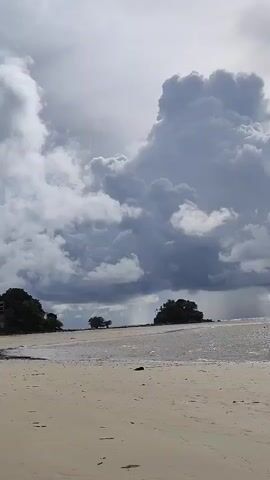 A static, vertical video shows a wide, sandy beach with a gentle wave breaking on the shore. In the distance, several small, tree-covered islands are visible. A large, dramatic cloud formation dominates the sky, with darker, rain-like streaks visible beneath it, suggesting impending weather. The water reflects the light, creating a shimmering effect. The scene is overcast with patches of bright sunlight breaking through. No people or other objects are clearly discernible in the video.