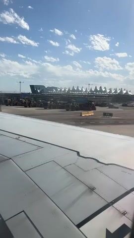 The video begins with a view from an airplane window. The foreground is dominated by the wing of the plane, showing its metallic surface with visible panel lines and rivets. Beyond the wing, a tarmac stretches out, with various construction vehicles, including yellow bulldozers and what appear to be other heavy machinery, parked in rows. In the background, the distinctive tent-like roofs of an airport terminal building are visible against a bright blue sky dotted with fluffy white clouds. The overall scene suggests the plane is either preparing for takeoff or has just landed at an airport under construction or undergoing maintenance.