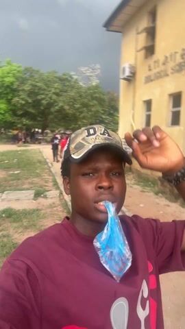 The video shows a young man wearing a maroon t-shirt and a camouflage baseball cap. He is walking outside, holding the brim of his cap and filming himself with a phone. He has a small blue plastic bag filled with liquid in his mouth. Other people are walking in the background. The sky is overcast. There is a building in the background with "Biology" visible on the side. At one point, he makes a surprised expression.
