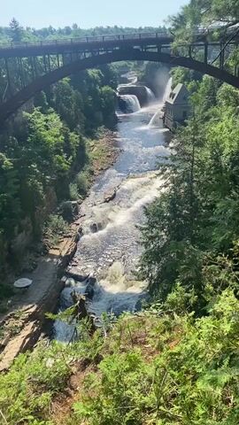 This video captures a scenic view of a river cascading over multiple waterfalls, viewed from a high vantage point. A large, dark metal bridge arches over the canyon, with a small, light-colored building situated on the right bank of the river, near the upper falls. The surrounding landscape is lush with dense green trees and vegetation, suggesting a natural park or wilderness area. The water is clear and flows rapidly, creating foamy white rapids and spray at the falls. The scene is bathed in bright sunlight, indicating it is daytime, likely a clear and warm day.

The primary subject is the natural landscape, dominated by the powerful flow of the river and its waterfalls, set within a verdant canyon. There are no visible people in the frame, and the only man-made objects of note are the prominent bridge and the small building. The activity taking place is the natural flow of water over the falls.

Notable details include the dramatic arch of the bridge, the contrast between the rugged rock formations of the canyon walls and the soft green foliage, and the presence of the building, which might be a historical structure or part of a hydroelectric facility. The overall impression is one of natural beauty and power, with a hint of human presence integrated into the landscape. There is no visible text in the media. The setting appears to be a location where a significant river flows through a gorge, creating a series of waterfalls.