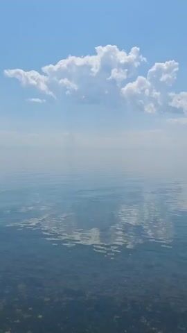 The video shows a calm, vast body of water under a clear blue sky with scattered white clouds. The water is transparent near the shore, revealing a rocky bottom. The surface of the water is mostly still, reflecting the sky and clouds. In the distance, a small boat with a dark hull is visible. The overall scene conveys tranquility and a sense of open space.