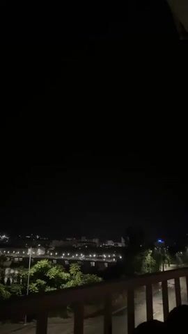The video shows a nighttime view of a city from a balcony. The city lights are visible in the distance and there are trees in the foreground. There is a bridge spanning a body of water. At one point, a flash of lightning illuminates the sky in a distinct purple hue.