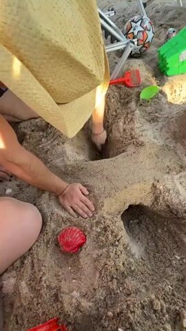 This media file shows a group of people at a beach, likely Nea Potidea, Greece. The scene is dominated by a large sand structure, possibly a sandcastle or a large hole being dug into the sand. A person wearing a straw hat is actively digging into the sand structure with their hands. Another person's arm is visible reaching into the sand. There are several beach toys scattered around, including a red shovel, a green lizard toy, and a red, shell-shaped object. The sun appears to be high in the sky, suggesting it's daytime, and the weather seems pleasant for a beach outing. The focus is on the collaborative effort of building or exploring the sand structure.
