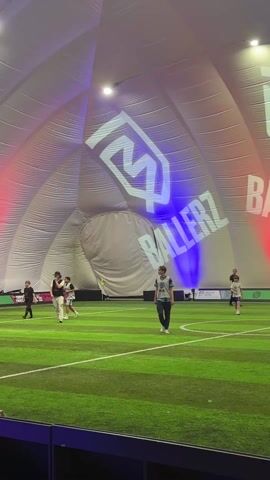 Several people are on an indoor soccer field with green artificial turf. The field is contained within a large dome structure. The dome’s inner surface is illuminated with red and blue lighting, and projects the white “BALLERZ” logo. People are walking, some are wearing soccer uniforms.