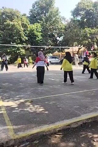 The video shows an outdoor volleyball game played on a paved court. Several women, most of whom are wearing hijabs and yellow shirts with black pants, are on the court. One person in the foreground is wearing a red shirt and white hijab with black pants. Another person in the background is wearing a white shirt and black pants. The game is in progress, with players in various stances, suggesting they are actively participating in the match. Trees and some buildings are visible in the background, indicating an outdoor setting, likely a school or community sports facility. The lighting suggests it is daytime.