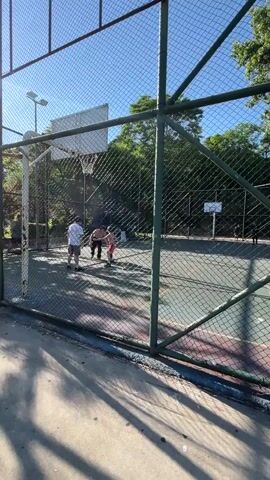 A group of people are playing basketball on an outdoor court. The court is enclosed by a chain-link fence. The sun is shining brightly, casting shadows on the ground. There are trees in the background. One person is shooting a basketball.