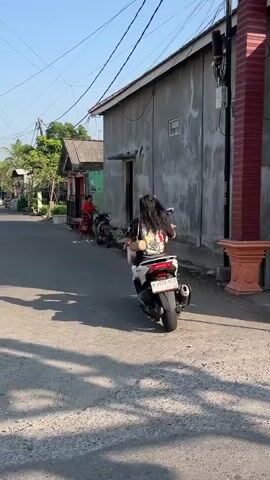 A person with long dark hair is riding a white scooter down a paved road. The scooter has a license plate that reads "N 4828 WCL". The person is wearing a white shirt with a graphic on the back and dark shorts. They are facing away from the camera. In the background, there are several buildings, including a gray one with a doorway and a smaller building with a red roof. There are also trees and power lines visible. Another person wearing a red shirt is standing near a motorcycle in the background. The sun is shining, casting shadows on the road.