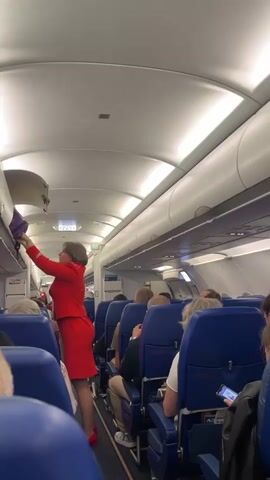 An overhead interior view of an airplane cabin shows a flight attendant in a red uniform closing an overhead storage bin. She is standing in the aisle, facing forward and to the left, with her right hand on the bin door. Several passengers are seated in blue seats, facing forward. The aisle is carpeted in dark gray. The ceiling of the cabin is white and illuminated by strip lights.