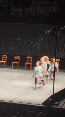 A stage performance is taking place. Four young women, dressed in similar light-colored shirts, grey skirts, black leggings, and brown shoes, are performing a dance routine on a stage. The stage has a glossy grey floor, and a dark backdrop with vertical crinkled patterns. Five wooden chairs are placed in a horizontal line towards the back of the stage. The women perform movements including bending over, shaking their hair, and walking/running. An audience is visible in the foreground, below the stage. Stage lighting is visible from the sides and above. The camera is stationary.