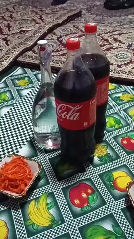 The camera, positioned at eye level, is focused on a low table covered with a patterned tablecloth. The tablecloth has a black and white checkerboard pattern interspersed with squares featuring illustrations of various fruits and vegetables. On the table, there are two large bottles of Coca-Cola, a clear glass bottle of water with a white stopper, and a small square dish filled with orange julienned carrots. The carrots appear to be prepared in a Korean-style. The table is set on a rug with a brown and beige pattern, adding warmth to the scene. In the background, partial views of people's feet wearing shoes and socks are visible, indicating the presence of individuals seated around the table. The scene appears to be a casual gathering or meal.