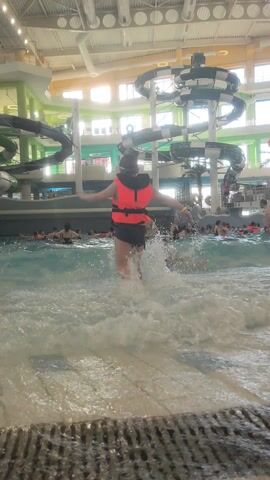 The video shows a waterpark with a wave pool filled with people. The pool has artificial waves, and children are playing in the water. A child in an orange life vest and black shorts is seen walking into the water. Another child in a pink swimsuit and pink arm floats walks toward the camera. In the background, there are large water slides with a twisting, turning structure. The waterpark has a high ceiling with exposed pipes. The scene is well-lit, with natural light coming through the windows.