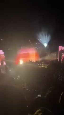 The video shows a band performing on a stage in front of a large crowd. The stage is lit with bright spotlights and colorful stage lights. The band consists of a singer in a black shirt and light jeans, a guitarist, a keyboard player, and a drummer. The singer is in the center of the stage, singing into a microphone and gesturing to the crowd. The guitarist is to the singer's right, playing an orange electric guitar. The keyboard player is in the background to the left. The drummer is mostly obscured in the background. The crowd is large and appears to be enjoying the performance.