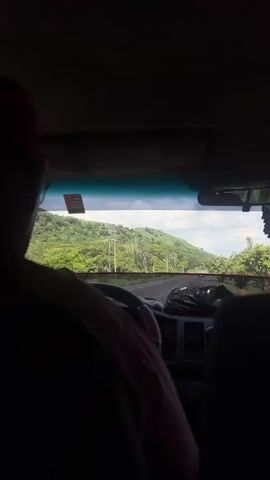 This media file shows the view from inside a vehicle, looking out through the windshield at a scenic road. The primary subjects are the road and the surrounding landscape, with a person's head and shoulder visible in the foreground, suggesting they are either driving or a passenger.

The scene is set on a paved road that winds through a lush, green, hilly terrain. The hills are covered in dense vegetation, indicating a tropical or subtropical environment. There are several utility poles visible along the side of the road, suggesting infrastructure in the area. The sky appears partly cloudy, with bright daylight, suggesting it is daytime. A black, tree-shaped air freshener hangs from the rearview mirror, a common object found in vehicles.

The activity is travel, with the vehicle moving along the road, as indicated by the perspective and the slight blur of the distant scenery. There are no strong emotional cues visible, but the overall impression is one of a journey through a beautiful natural landscape.

Visible text includes a sticker on the windshield, which appears to be an inspection or registration tag. The text is not clearly legible due to the resolution and angle. Another sticker, possibly a warning or information label, is present on the interior of the vehicle, above the windshield. A small rectangular object, possibly a parking permit or toll sticker, is also visible on the windshield. The location context provided is San Lorenzo, Honduras.