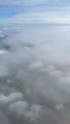 This is an aerial view of clouds from an airplane. The camera is angled downwards, showing a vast expanse of white and gray clouds. Patches of land and water are visible beneath the clouds in places. The sky above is blue with some scattered white clouds. The video appears to be a continuous shot, with no discernible actions or interactions occurring within the frame. The environment is that of the atmosphere from an elevated perspective.