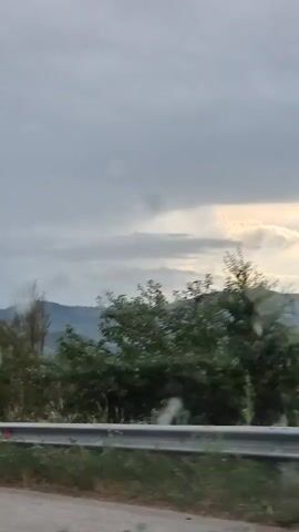 This media file captures a scenic view from a vehicle traveling along a road in Osikovska Lakavitsa, Bulgaria. The scene is dominated by rolling hills covered in lush green trees, suggesting a rural or mountainous landscape. In the distance, several houses are visible, nestled amongst the trees, indicating a small village or settlement. The sky is overcast with gray clouds, hinting at cloudy or possibly rainy weather, though some patches of lighter sky suggest that the sun might be trying to break through. The time of day appears to be late afternoon or early evening, given the subdued light. A metal guardrail runs along the side of the road in the foreground, and the road surface itself is visible, appearing wet, which could be from recent rain. There are several blurry spots or smudges on the lens through which the video is filmed, possibly water droplets or dirt. No people are clearly visible, and the focus is on the natural landscape and the passing scenery. The overall mood is peaceful and contemplative, characteristic of a road trip through the countryside.