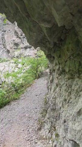 The video shows a narrow, gravelly path nestled alongside a sheer rock face in an outdoor setting. The path is overshadowed by an overhanging rock formation. Green foliage and small trees dot the landscape on one side of the path. Further along the path, two individuals, one in blue and the other in red, can be seen walking away from the camera.