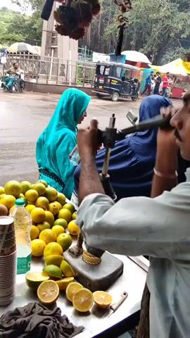 A street vendor is making juice from oranges and limes. He is using a hand-cranked juicer. There are piles of oranges and limes on the cart. Two women are standing in the background, one wearing a blue headscarf and the other a turquoise one. An auto-rickshaw is visible behind them. The scene appears to be outdoors, with a tree and a building in the background.