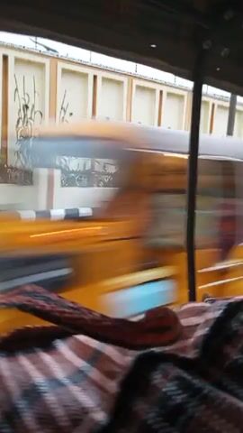The video shows a view from inside a vehicle, looking out the window. The window has a green tint. In the foreground, a patterned fabric, possibly a blanket or seat cover, is visible. Outside the window, a road is seen with a black and white striped curb. Beyond the curb is a wall with regularly spaced vertical elements and horizontal trim. Green foliage is visible behind the wall. The road surface appears wet in places, suggesting it may have rained. The vehicle appears to be in motion, as indicated by the blur of the passing scenery. There are no people clearly visible in the frame.