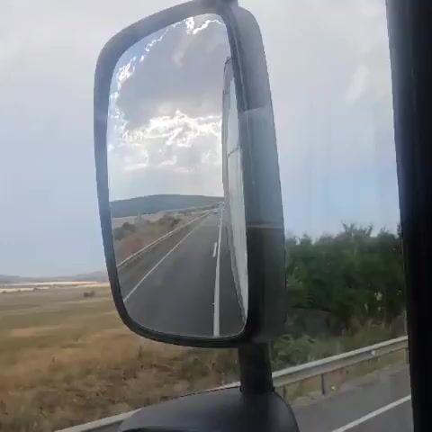 The video shows a view from a truck's side mirror, reflecting a highway with a cloudy sky overhead. The truck is in motion, indicated by the blurred surroundings and the continuous road ahead. The reflection captures the highway stretching into the distance, bordered by a guardrail and dry vegetation. Hills and trees are visible on either side of the road. The sky is filled with dramatic, sunlit clouds. The audio consists of a person speaking in Bulgarian, expressing a desire for everyone to be happy and to be better towards each other.